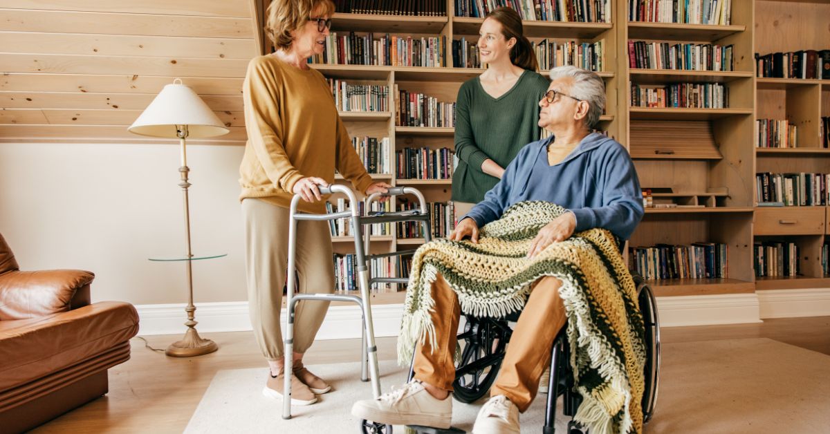 Caregiver supporting elderly parents at home – woman using a walker, man in wheelchair covered with blanket, and adult daughter offering assistance in a cozy room with bookshelves.