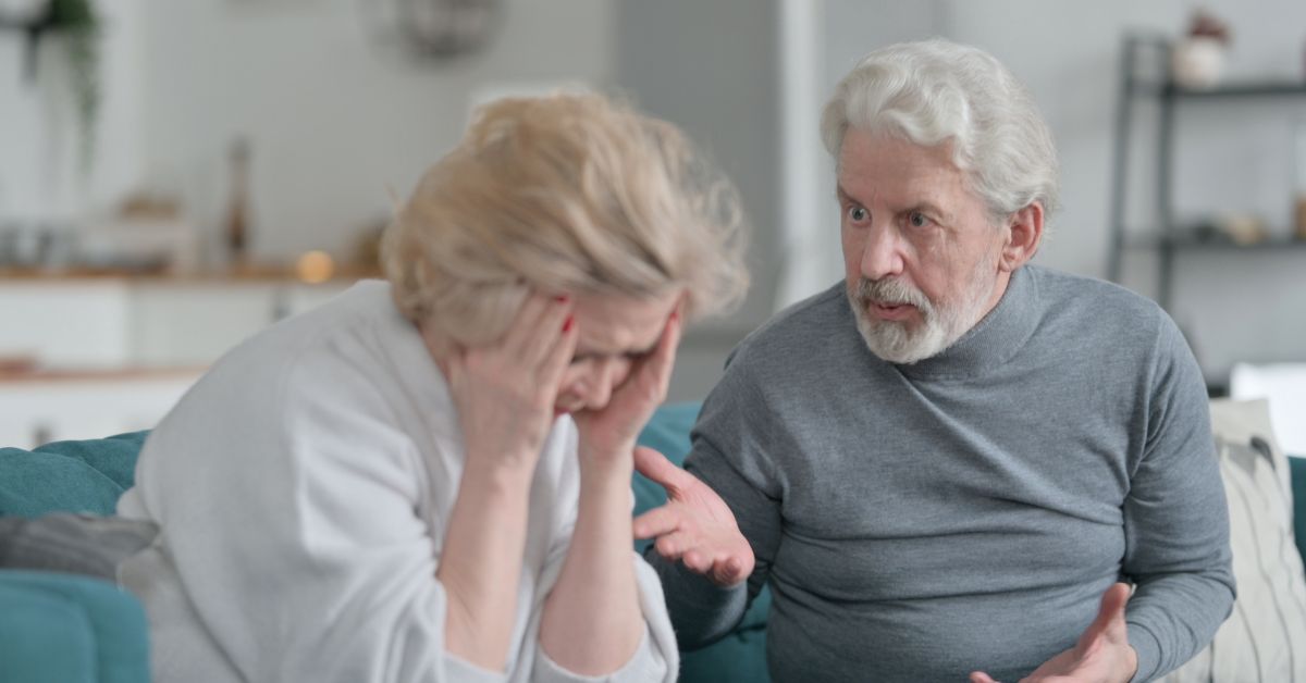Senior couple having an emotional argument at home, with one partner looking frustrated and the other holding their head in distress.