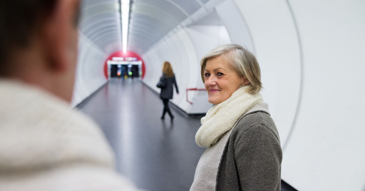 Mature woman smiling in a modern tunnel walkway, turning toward a person in the foreground, suggesting a meaningful or emotional farewell moment.