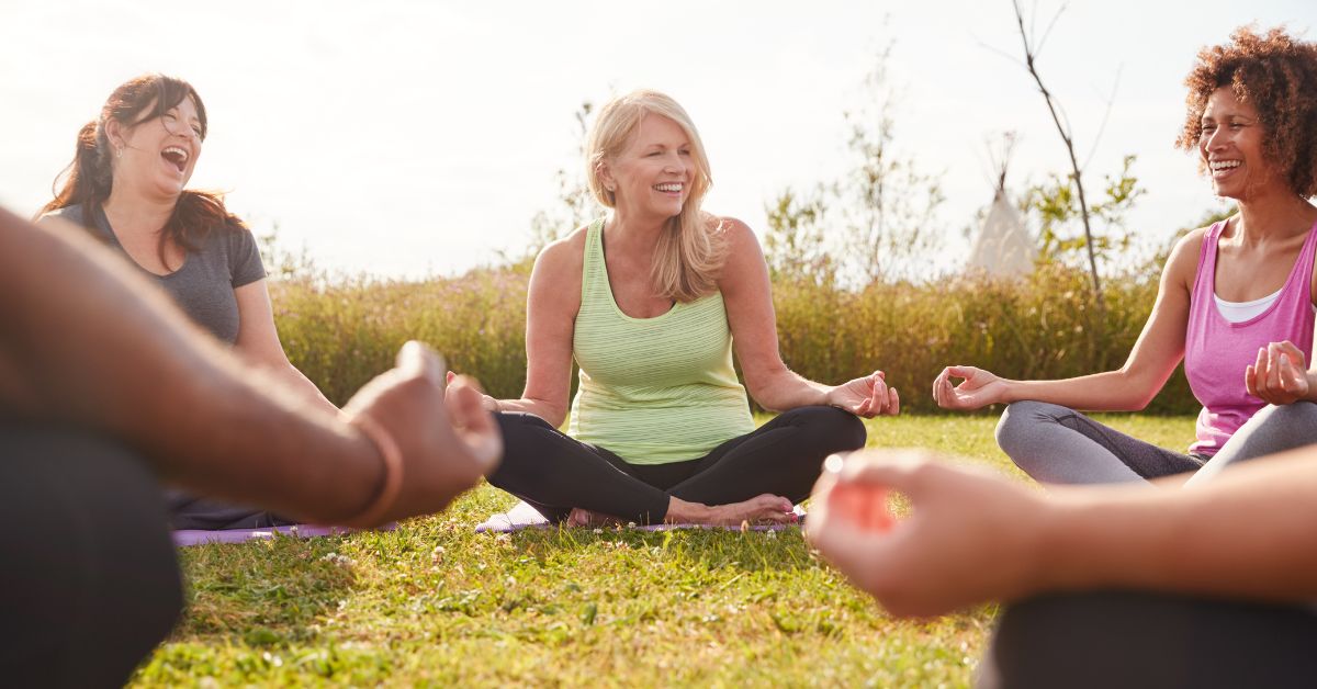 Group of mature women laughing and practicing outdoor meditation together on yoga mats in a grassy field, promoting mindfulness, happiness, and well-being in later life.