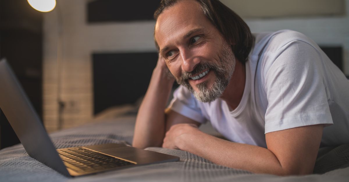 Middle-aged man smiling while lying on bed and video chatting on a laptop during an online conversation.