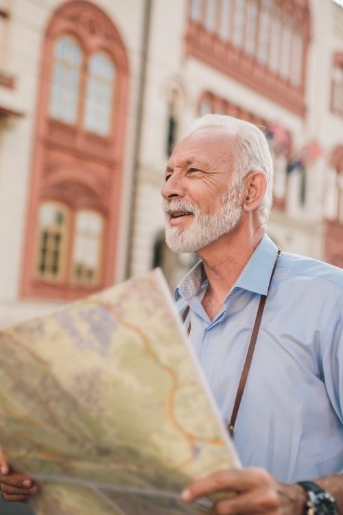 Smiling mature man with a white beard holding a large map while exploring a historic city, wearing a light blue shirt and a camera strap around his neck.