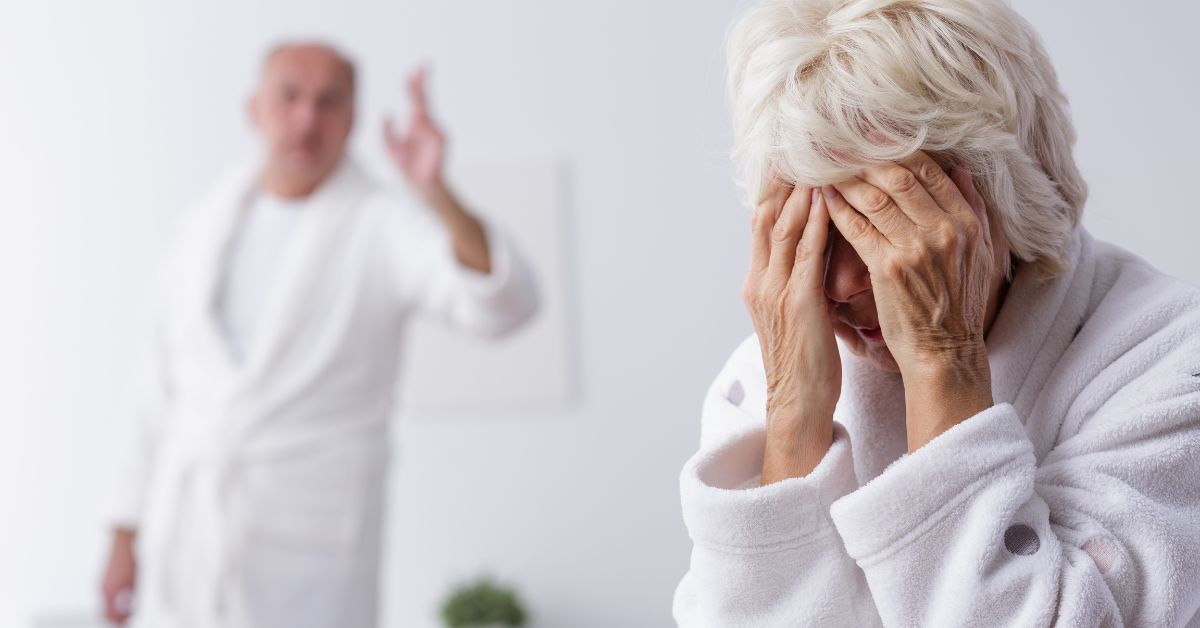 Upset senior woman covering her face while an older man in the background appears to be arguing, representing emotional distress in a toxic relationship.