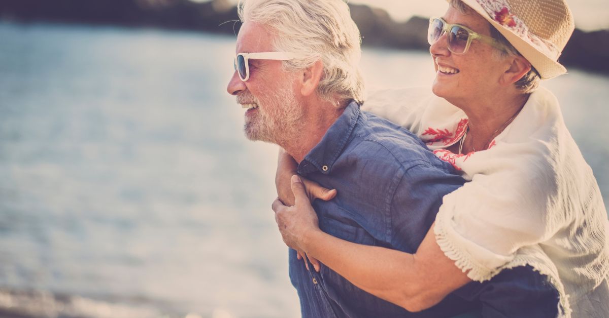 Happy senior couple enjoying a playful piggyback ride at the beach, symbolizing love, joy, and connection in later life relationships.