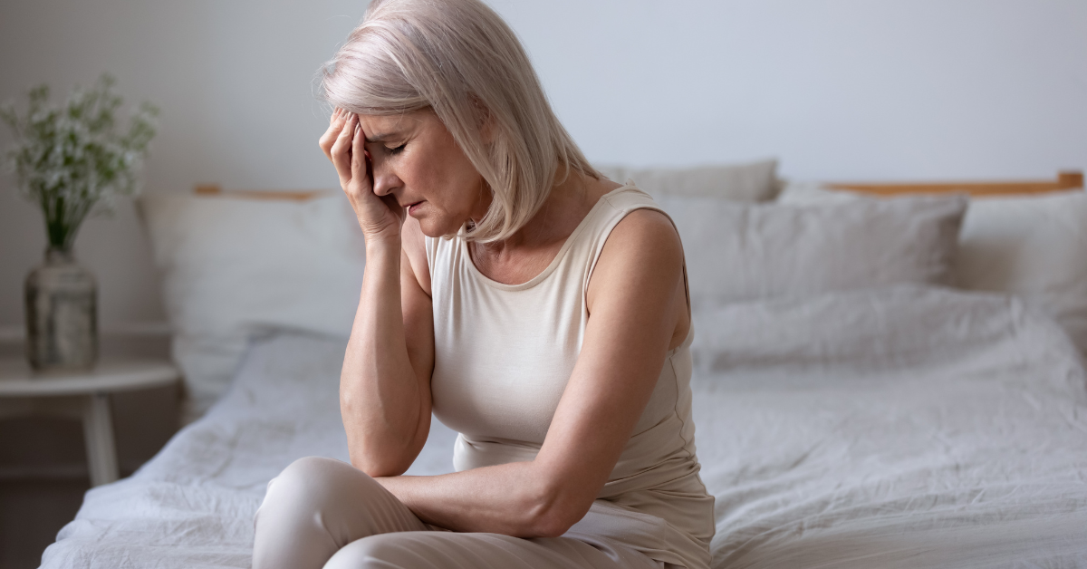Middle-aged woman sitting on bed with hand on forehead, showing signs of menopause symptoms like fatigue, stress, or hot flashes.