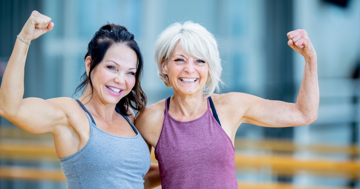 Two fit women over 50 smiling and flexing their arms in workout clothes, showing strength, confidence, and healthy aging through fitness.