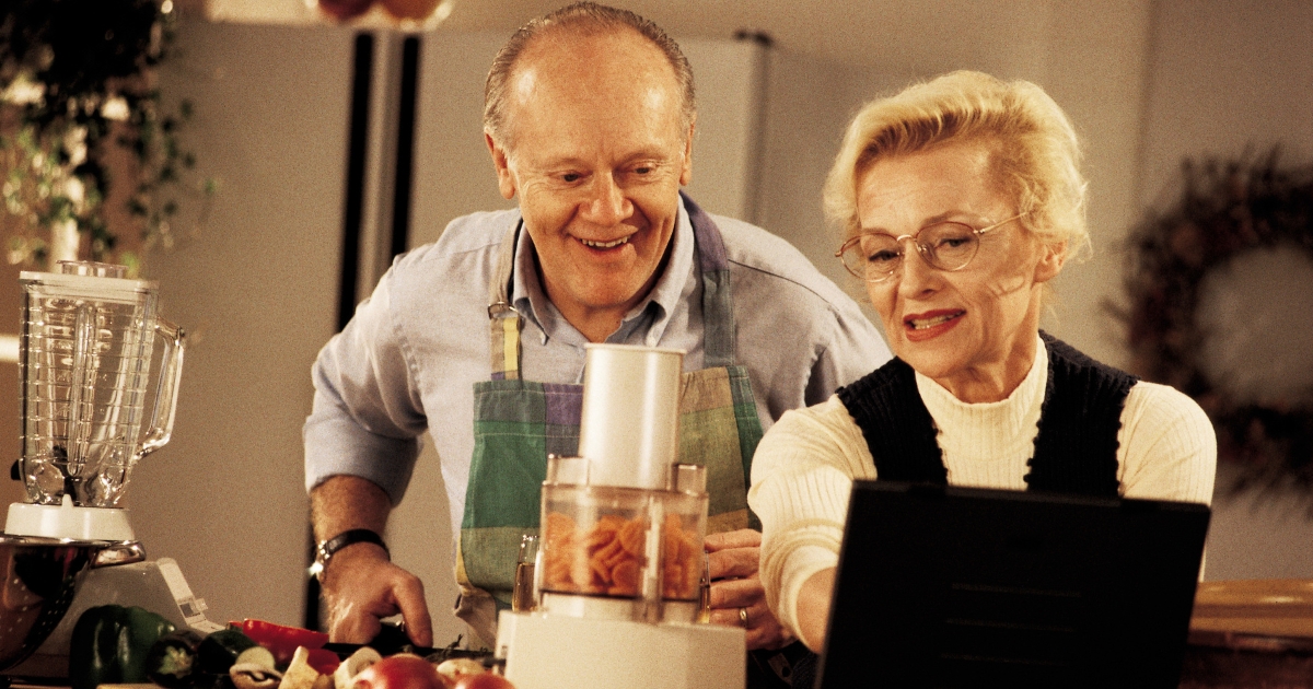 Smiling senior couple cooking together in the kitchen, using fresh vegetables and a food processor, enjoying a shared hobby.