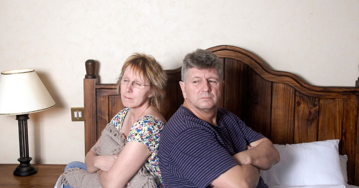 Middle-aged couple sitting in bed back-to-back with arms crossed, appearing emotionally distant and frustrated — symbolizing conflict or disconnection in a sexless relationship.