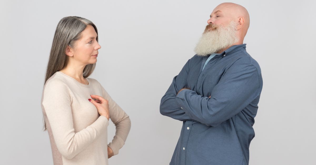 Mature couple facing each other with serious expressions, arms crossed – tension and reflection in a relationship conversation.