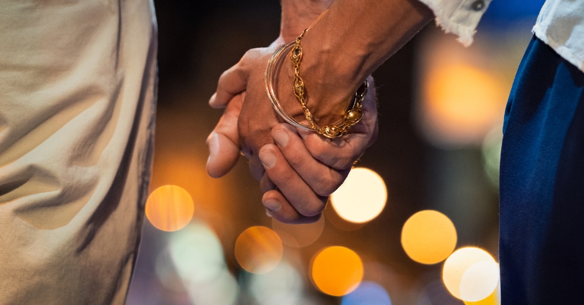 Staying Safe When Online Dating In Your 50s And Beyond - Mature Singles Finding Love Close-up of two people holding hands at night, with a blurred cityscape and glowing lights in the background. One person wears gold bracelets, symbolizing connection, romance, and companionship.