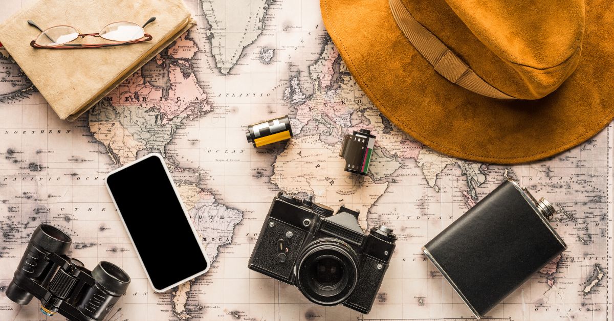 The Art of Solo Travel for Mature Adventurers - Mature Singles Finding Love Flat lay of vintage travel essentials on a world map, including a camera, smartphone, film rolls, travel journal with glasses, binoculars, a hip flask, and a brown suede hat.