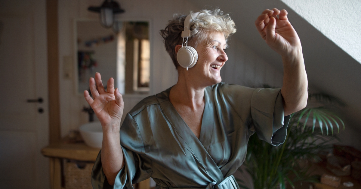 The Role of Self-Love in Strengthening Relationships After 50 - Mature Singles Finding Love Joyful senior woman dancing and listening to music with headphones in a cozy home, embracing self-love and confidence after 50.