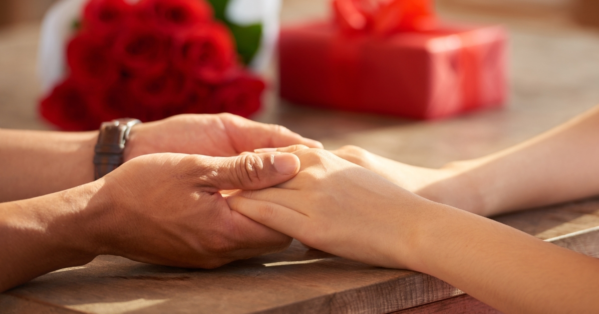 Top 5 Things Men Look For In A Soulmate - Mature Singles Finding Love Close-up of a couple holding hands across a wooden table, with a romantic background featuring red roses and a gift box, symbolizing love and connection.