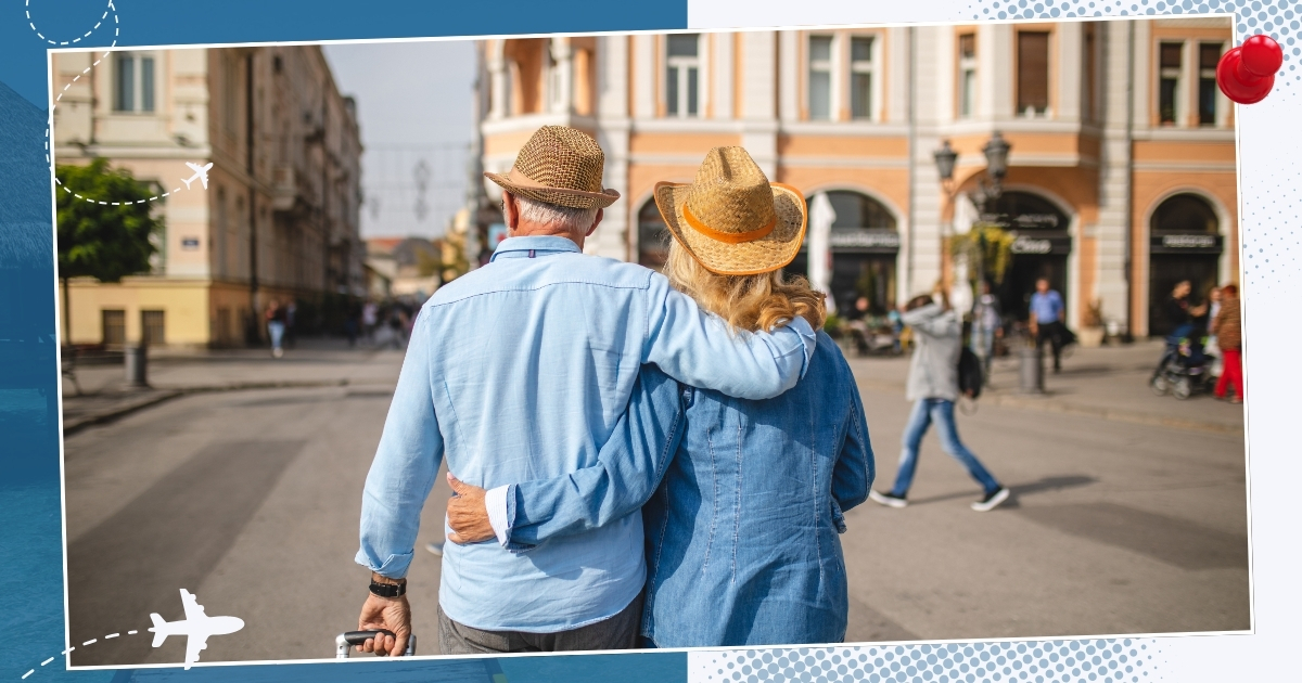 Older couple in matching denim outfits and straw hats walking arm in arm through a European-style city square, symbolizing travel and romantic getaways for mature adults.