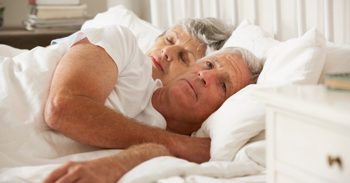 Senior couple in bed, man looking thoughtful while partner sleeps