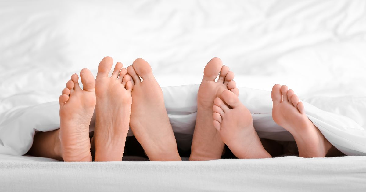 What If Your Partner Wants A Threesome - Mature Singles Finding Love Three pairs of feet under white bedsheets, suggesting intimacy and relationships.
