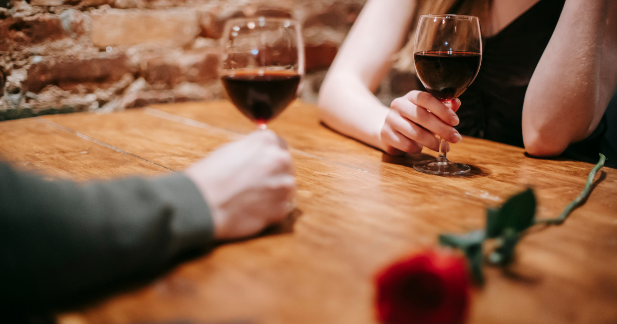 What To Talk About On A First Date - Mature Singles Finding Love Close-up of two people holding glasses of red wine at a wooden table with a single red rose, suggesting a romantic first date setting.