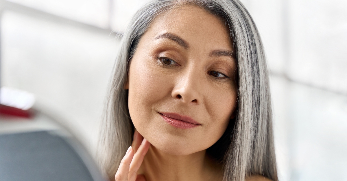 What You Should Know About Dating An Older Woman - Mature Singles Finding Love A confident older woman with long silver hair looking at her reflection in a mirror, gently touching her neck. She has a calm and content expression, with natural beauty and grace.