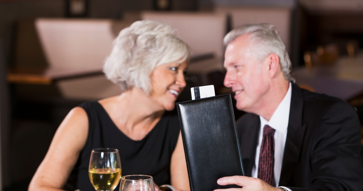 Who Should Pay On A First Date - Mature Singles Finding Love Smiling mature couple sitting at a restaurant table with wine glasses, as the man holds a black check holder with a receipt inside.