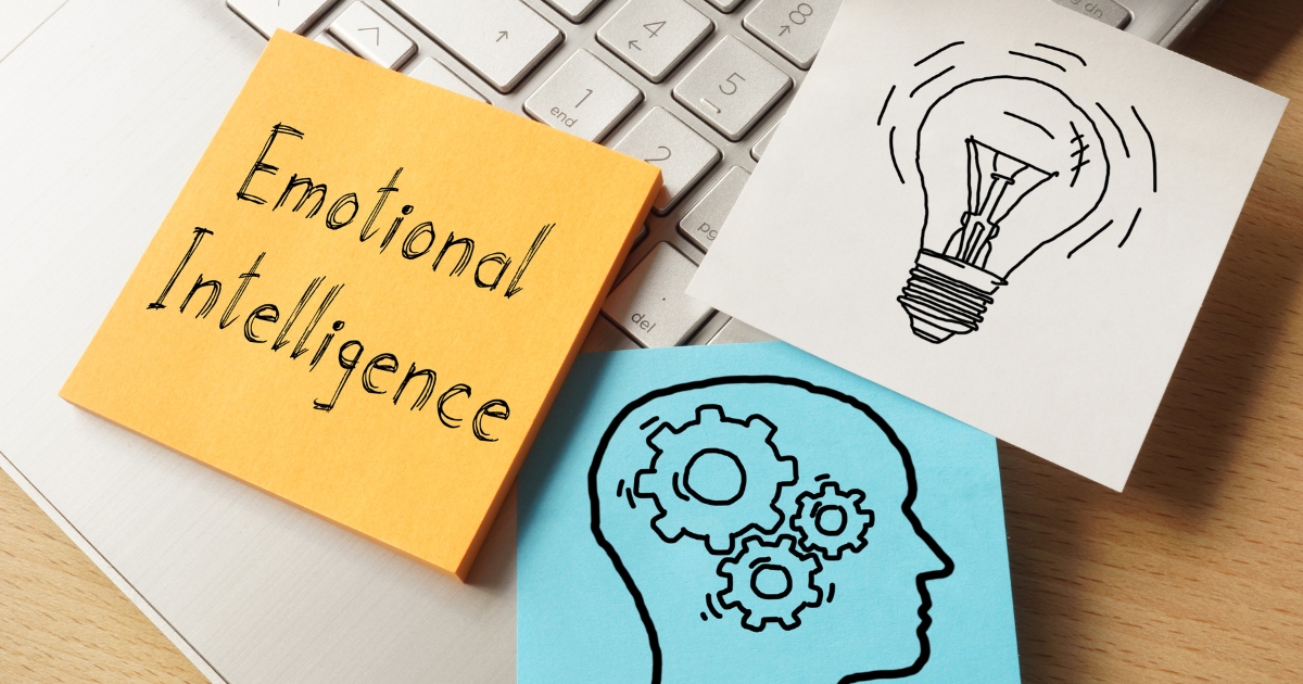 Sticky notes on a keyboard showing the words 'Emotional Intelligence' and illustrations of a lightbulb and a head with gears, symbolizing self-awareness and insight.